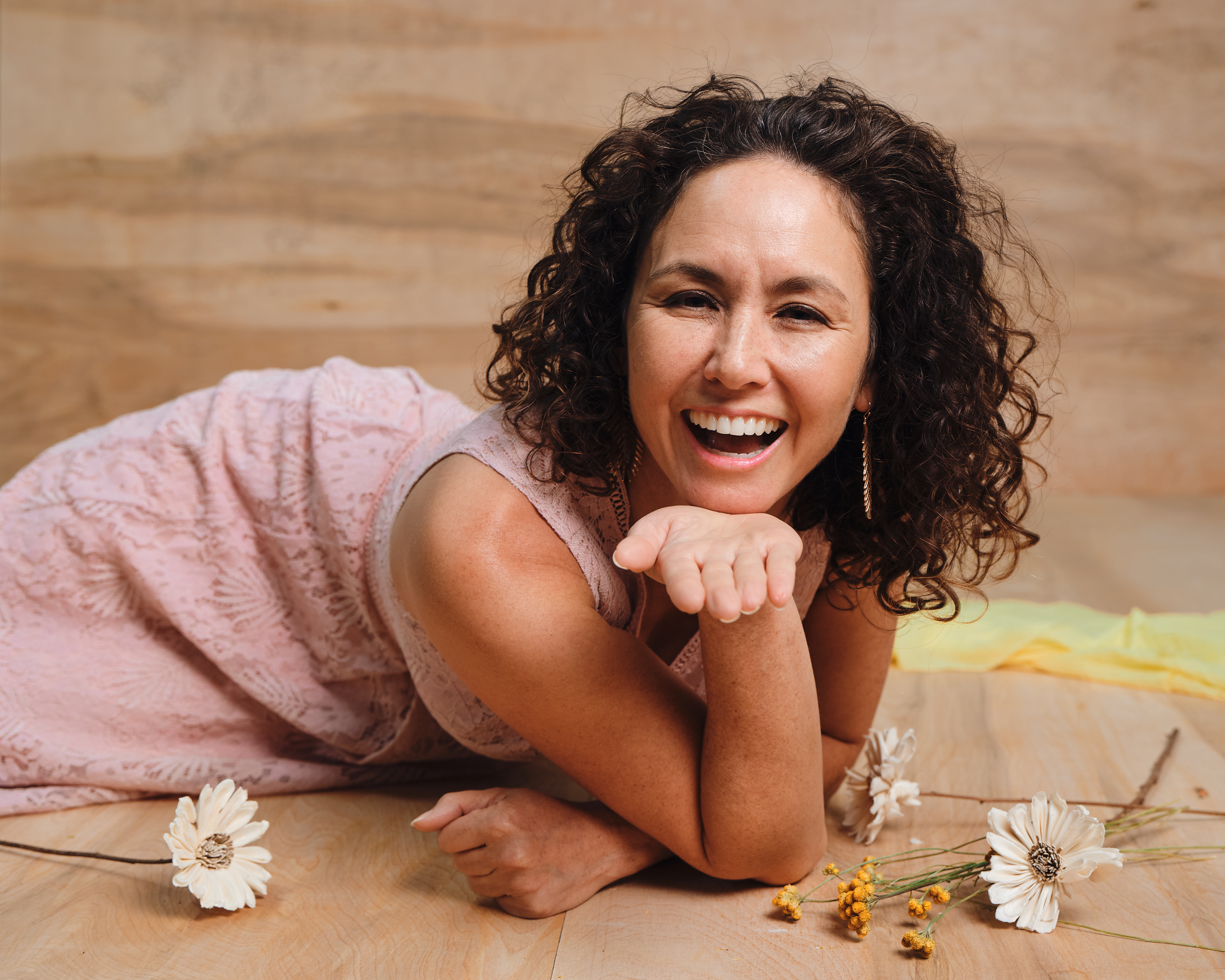 A woman with curly dark hair, wearing a pink dress, lies on a wooden floor surrounded by flowers.
